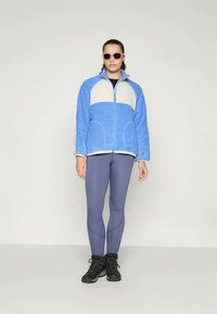 Sinine fliisdžemper valgete paneelidega, lukuga kinnitamine, koos hallide retuuside ja mustade saapatega. Modelli peal on prillid.