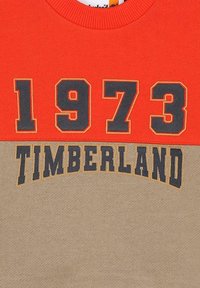 Maglione rosso e beige con grandi numeri stampati "1973" e la parola "TIMBERLAND" in nero. Collo a costine, materiale in cotone.