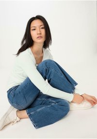 Maglietta a maniche lunghe verde chiaro, pantaloni in denim blu larghi e sneaker bianche. Modello seduto, gambe piegate, con le mani sui piedi, sfondo neutro.