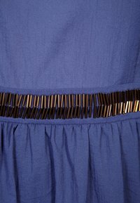 Sininen teksturoidulla kankaalla valmistettu mekko, jossa on gathered-hame ja vaakasuora metallikoriste hihassa.