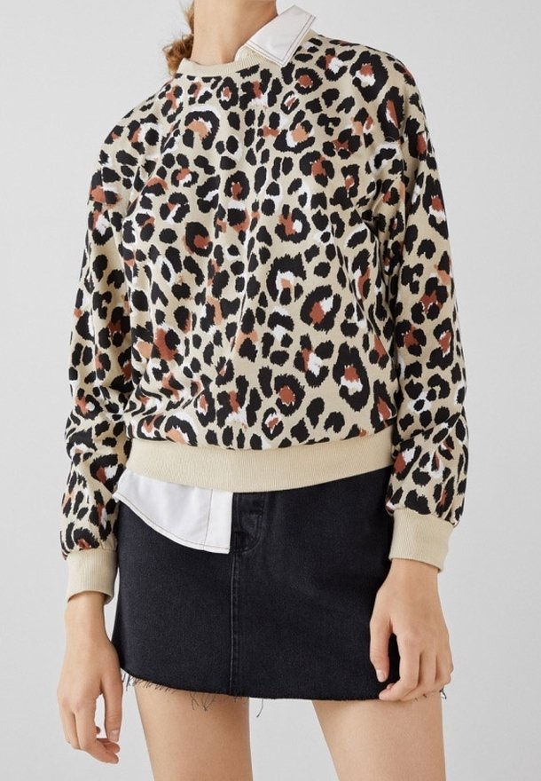 Femme portant un pull beige et marron à motif léopard sur une chemise blanche, associé à une mini-jupe en jean noire.