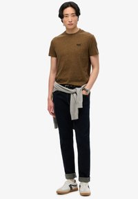 Camiseta corta de manga larga texturizada marrón, vaqueros de mezclilla oscuros, suéter gris claro atado a la cintura, zapatillas blancas con detalles negros, reloj en la muñeca.