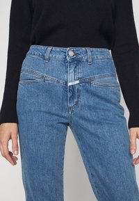 Jeans de mezclilla de cintura alta en azul claro con cierre de cremallera, cinco bolsillos y costuras en contraste, con una textura suave y un diseño minimalista.