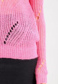 Gestrickter Pullover in leuchtendem Pink, mit offenen Strickmustern und Ausschnittdetails an den Ärmeln. Weiche Textur mit gerippten Bündchen.