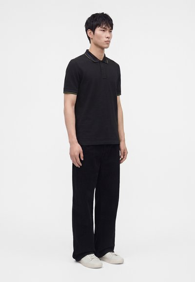 Jeune homme debout, portant un polo noir avec col et manches rayés, pantalon noir et baskets blanches sur un fond blanc uni.