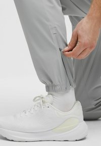 Pilki sportiniai kelnės su elastingomis manžetėmis ir šoninėmis užtrauktukais. Asmuo reguliuoja užtrauktuką, vilkėdamas baltus sportbačius su tekstūruota tinkleliu.