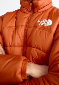 Orange puffer jacket with a high collar and front zip. Features quilted texture and a white logo on the chest. Elastic cuffs and hem.