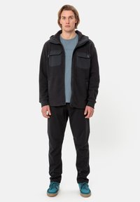 Giacca in pile grigio scuro con cappuccio, due tasche sul petto e cerniera. Indossata sopra una camicia azzurro chiaro e abbinata a pantaloni neri e scarpe turchesi.