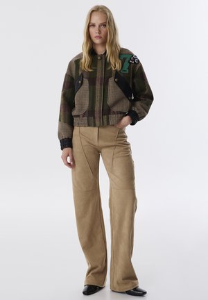 Cazadora bomber a cuadros en tonos verdes y marrones, con detalles en negro en los bolsillos y un parche decorativo. Pantalones de pierna ancha beige.