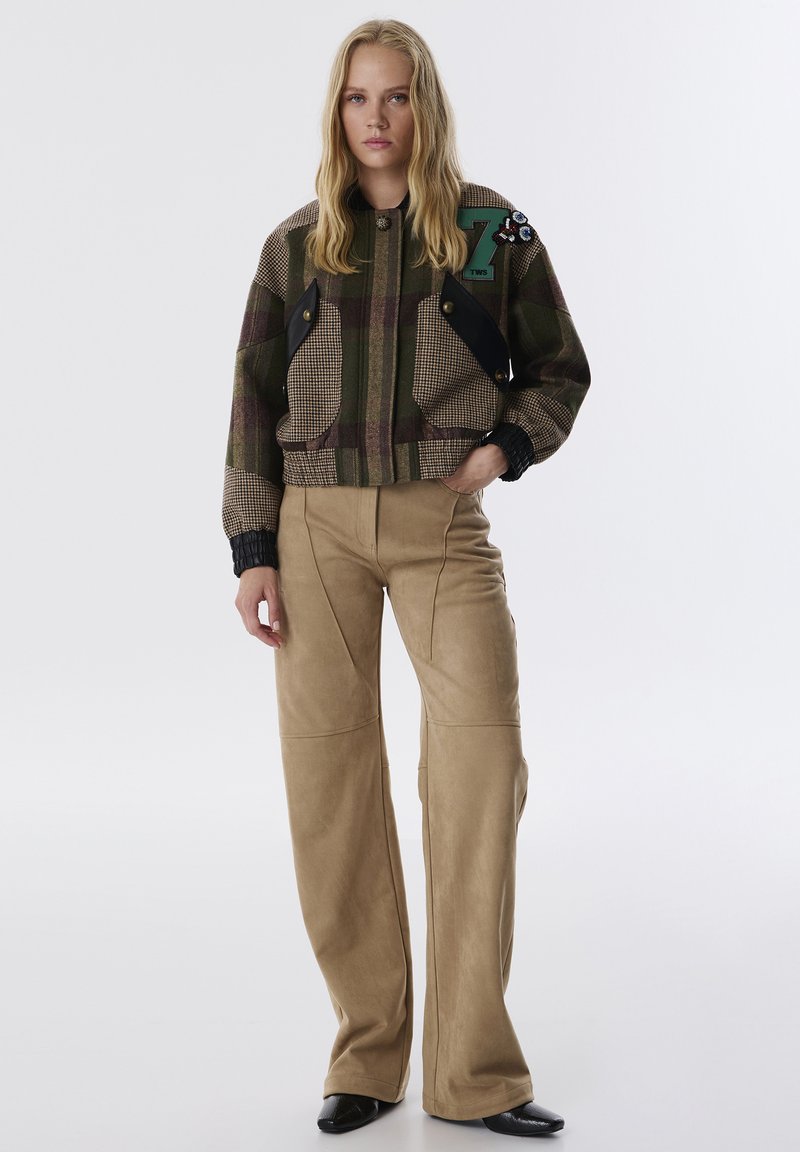 Afgekorte geruite bomberjack in tinten van groen en bruin, met zwarte zakaccenten en een decoratieve patch. Beige wijde broek.