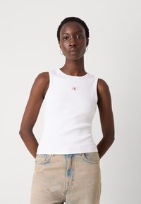 Femeie stând cu mâinile la spate poartă un top alb fără mâneci cu un logo mic și blugi deschiși la culoare, pe un fundal simplu.