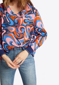 Kolorowa, oversized bluzka z abstrakcyjnymi wirami w odcieniach różu, niebieskiego i pomarańczowego. Projekt z dekoltem w kształcie litery V, z mankietami i miękką tkaniną.