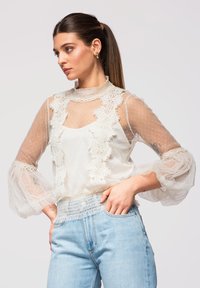 Femme avec queue de cheval brune portant un chemisier en dentelle blanche transparente et un jean taille haute bleu clair, regardant vers la gauche avec les mains sur les hanches.