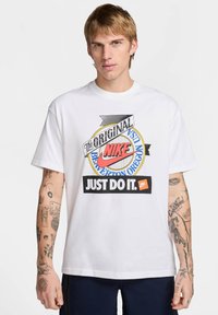 Camiseta de algodón blanca con un estampado gráfico colorido que incluye el texto "Nike", "Just Do It" y "Beaverton Oregon". Mangas cortas, cuello redondo.
