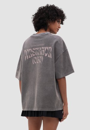 Osoba s kudrnatými vlasy, která nosí šedé oversize tričko s růžovým nápisem "CRAFTING AESTHETICS WRSTBGR (030)" na zádech, doplněné černou sukní.