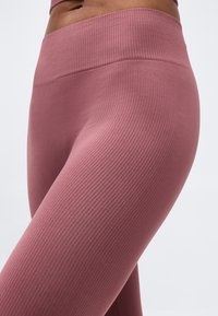 Κοντινό πλάνο ενός ατόμου που φοράει ψηλόμεσα, ριβδωτά, μωβ κολάν με αόρατη μέση, σε απλό φόντο.