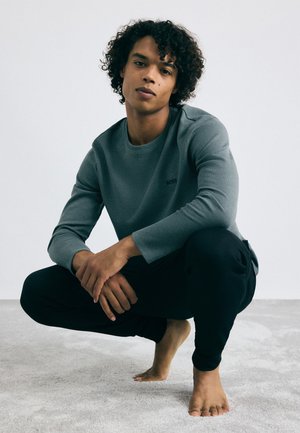 Graues, strukturiertes Langarmshirt mit rundem Ausschnitt und schwarzem Logo, kombiniert mit schwarzen Hosen. Das Model posiert barfuß auf einer hellgrauen Oberfläche.