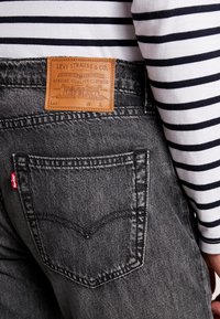 Jean en denim gris foncé avec une étiquette en cuir portant la mention "Levi Strauss & Co." et une étiquette rouge sur la poche arrière. Une chemise à rayures à manches longues est visible.