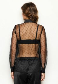 Schwarze Bluse aus durchsichtigem Stoff mit Polka-Dots, Kragen, langen puffigen Ärmeln, elastischem Bund und schwarzen Stoffakzenten an Schultern und Manschetten.
