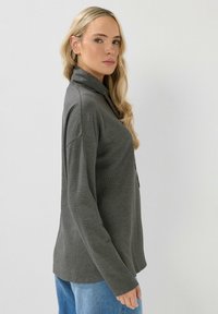 Harmaa ribattu pitkähihaisessa paidassa on kauluskainalo ja nappiyksityiskohta. Yhdistetty sinisiin farkkuihin, esitetty neutraalissa ympäristössä.