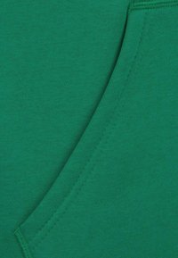 Zelená látka mikiny s hladkou strukturou a šikmým švem. Obsahuje žebrované okraje a kapsu s prošívanými detaily.