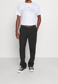 White tie-dye t-shirt with "WELLNESS" text, paired with black trousers and navy sneakers. Black belt with silver accents completes the outfit.