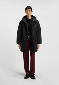 Cappotto imbottito nero con cappuccio, dotato di tasche laterali, zip frontale e una texture liscia. Indossato sopra una maglietta scura e pantaloni borgogna.