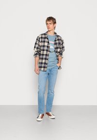 Jovem de pé com as mãos nos bolsos, vestindo uma t-shirt gráfica azul, uma camisa xadrez azul marinho e branca, jeans claros e ténis de duas cores.