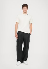 Hvit t-skjorte med korte ermer og logo, kombinert med svarte cargo-bukser med sidelommer. Hvite sneakers fullfører antrekket.