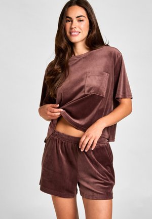 Femme aux longs cheveux bruns portant un short en velours marron ample et un haut à manches courtes, tenant l'ourlet de sa chemise et souriant devant un fond uni.