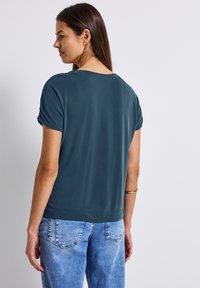 Tyrkysový top s krátkým rukávem a volným střihem, s ohrnutými rukávy, v kombinaci s světle modrými džíny. Hladká textura, jednoduchý design, bez vzorů.
