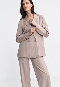 Beżowy garnitur w prążki z podwójnym zapięciem marynarki i szerokimi nogawkami spodni. Gładka faktura materiału, z brązowymi guzikami i klapami.