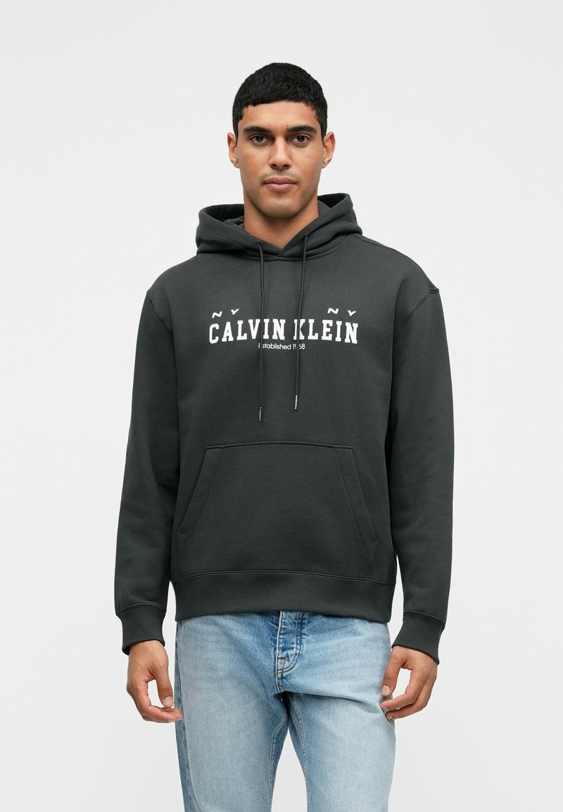 Hoodie verde escuro com bolso tipo canguru e cordões, apresentando o texto logo em branco "Calvin Klein". Textura suave, corte descontraído.