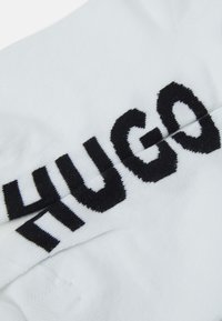Fehér textil, amelyen egy feltűnő fekete "HUGO" logó található. Az anyag finom textúrájú, látható varrási részletekkel.