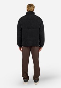 Giacca puffer in corduroy nera con colletto alto, sezioni trapuntate e polsini a coste, abbinata a pantaloni marroni e sneakers nere.