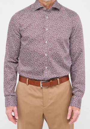 Mann trägt ein langärmeliges, gemustertes Hemd, das in beige Hose mit braunem Ledergürtel gesteckt ist.
