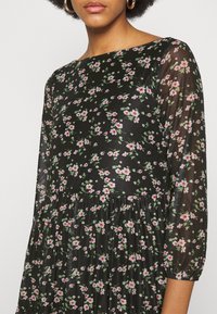 Robe noire à fleurs avec des manches longues transparentes, présentant des motifs de fleurs rouges et roses, une taille froncée et un col rond.