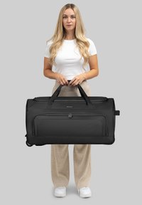 Grand sac de sport noir en tissu durable, doté de deux poignées supérieures, d'une poche frontale zippée et de roulettes lisses pour un transport facile.