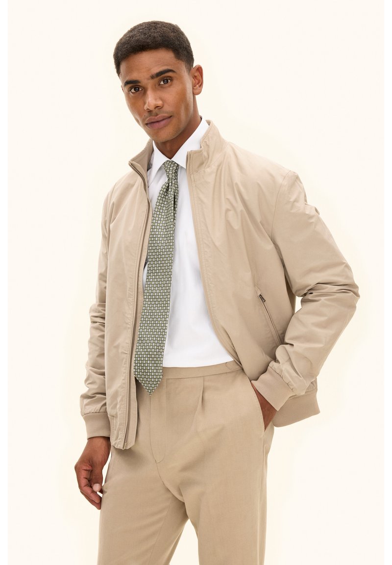 Beige Bomberjacke aus leichtem, glattem Stoff mit Stehkragen und gerippten Bündchen, kombiniert mit einer strukturierten grünen Krawatte und einer Anzughose.