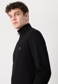 Svart stickad zip-up jacka med hög krage, med en texturerad tyg och en liten logotyp på sidan.