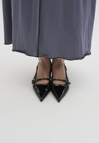 Schwarze Lackleder-Pumps mit spitzem Zeh und Knöchelriemen. Sie haben eine glänzende Textur und eine schlanke Silhouette und befinden sich unter einem fließenden grauen Kleidungsstück.