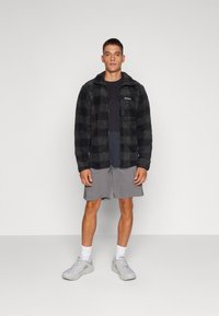 Giacca in pile con motivo a scacchi neri, dotata di zip, tasca sul petto, abbinata a una t-shirt scura e pantaloni grigi, indossata con scarpe da ginnastica.