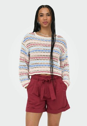 Kvinde med langt flettet hår iført en stribet åbenstrikket sweater og vinrøde højtaljede shorts med bindebånd, med hænderne i lommerne.