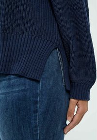 Marineblauer Strickpullover mit geripptem Saum und seitlichen Schlitzen, kombiniert mit blauen Jeans. Die Struktur wirkt dick und gemütlich mit einer lockeren Passform.