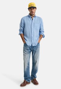 Camisa de botones de color azul claro, jeans de mezclilla desgastados, zapatos marrón con cordones y una gorra amarilla con acentos en azul marino. Presenta una bufanda anudada en el cuello.