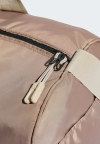 Sac de sport en nylon rose clair avec une fermeture éclair noire et deux tirettes blanches, complété par une sangle beige et des accents texturés.