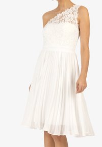 Bílé šaty s jedním krajkovým ramínkem, plisovanou sukní, přiléhavým korzetem a texturovanými květinovými krajkovými detaily. Hladká, lehká tkanina.