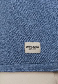 Materialul unui pulover tricotat albastru, prezentând un prim-plan al unui etichetă de culoare bej deschis pe care este imprimat cu negru "JACK&JONES EST.1990". Textură medie, cu greutate medie.