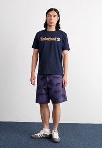 Jūras spēku kokvilnas T-krekls ar "Timberland" logo, saskaņots ar purpura krāsas raibajiem šortiem. Modelis valkā pelēkus un baltus sporta apavus.