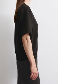 Černá mikina s krátkými širokými rukávy a zaobleným výstřihem. Měkká, texturovaná tkanina a přiléhavý spodní lem. Boční pohled.